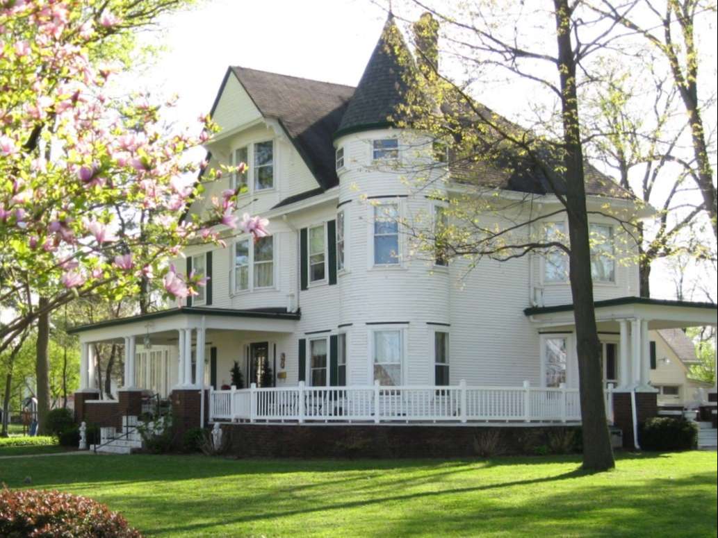 white three story late Victorian residence with wraparound porch, green lawn, flowering trees